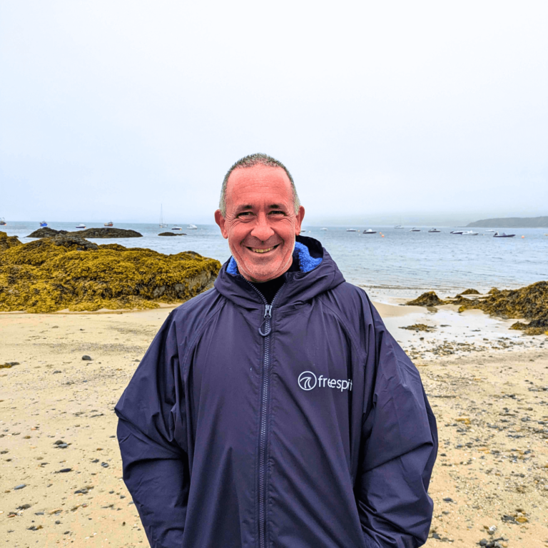 Bob is standing on Porthdinllaen beach wearing a dryrobe. He has his hands in his pockets.