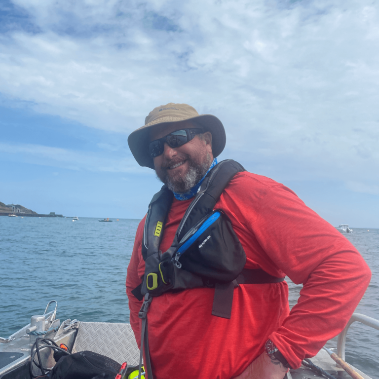 Neil is standing on board Project Seagrass' boat Gwen. He is wearing a hat, sunglasses, and a red coat.