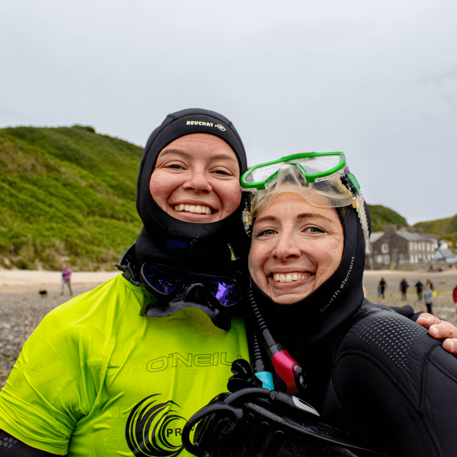 Hannah is wearing a yellow Project Seagrass rash vest and has her arm around volunteer Antje following a snorkel session to collect seagrass. They are on Porthdinllaen beach.