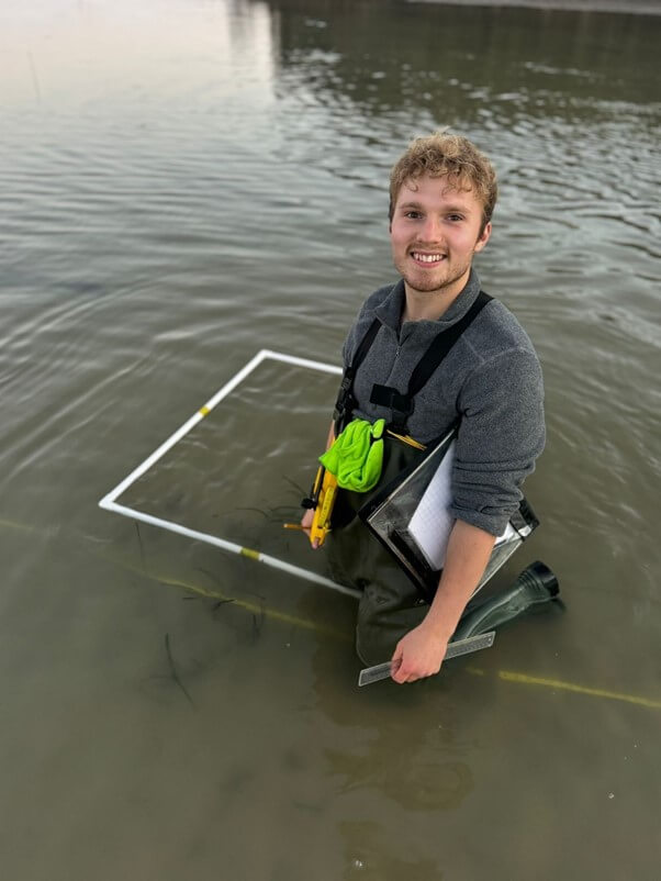 Finn (one of Project Seagrass' 2024-25 interns) is wearing waders and kneeling in the water. He is kneeling next to a quadrat along a transect line. He is holding a ruler and pencil and has a folder with monitoring sheets tucked under his arm.