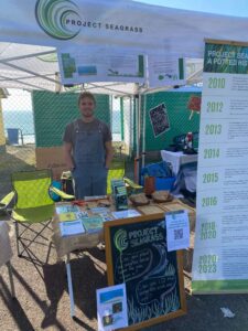 Project Seagrass 2024-25 Intern Finn is standing behind the Project Seagrass stand at the Toxic Trophy in West Wales. He is wearing a t-shirt and dungarees and has his hands in his pockets. On the stand as a range of leaflets, stickers, and postcards. To the side of the stall is a banner outlining Project Seagrass' history.