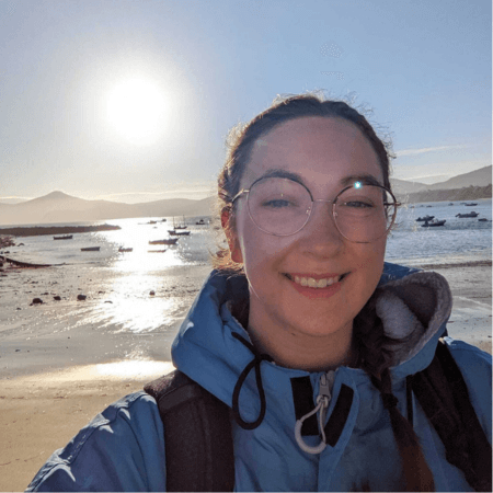 Emma is standing on the beach with the sun in the background. She is wearing a blue coat.