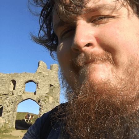 Adam is standing by a castle ruin. He has a beard and moustache.