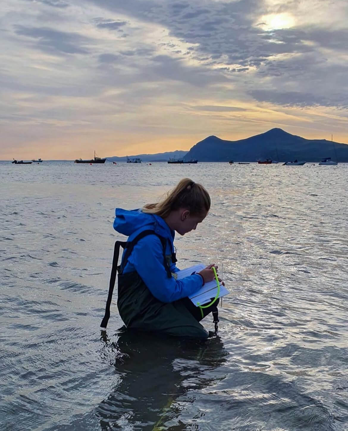 Lowri is crouched in the shallow water at Porthdinllaen. She is wearing waders and making monitoring notes.