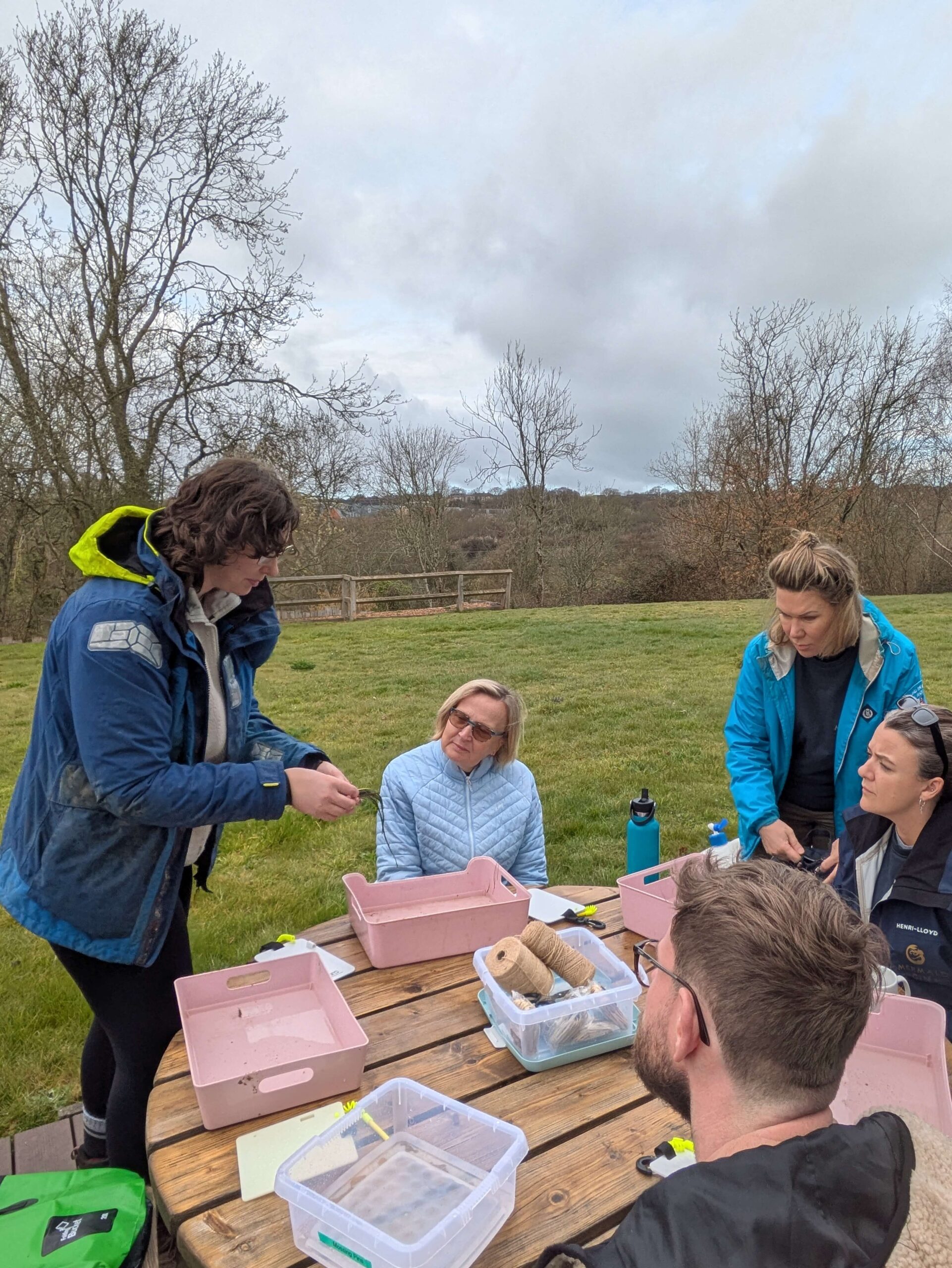 Emma is showing volunteers how to prepare transplants at a workshop at Liz Earle on the Isle of Wight. She is wearing a blue jacket and is standing next to a picnic bench where a group of volunteers are gathered.