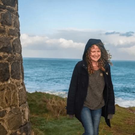 Laura is walking along a grassy path by the coast. She has her hood up.