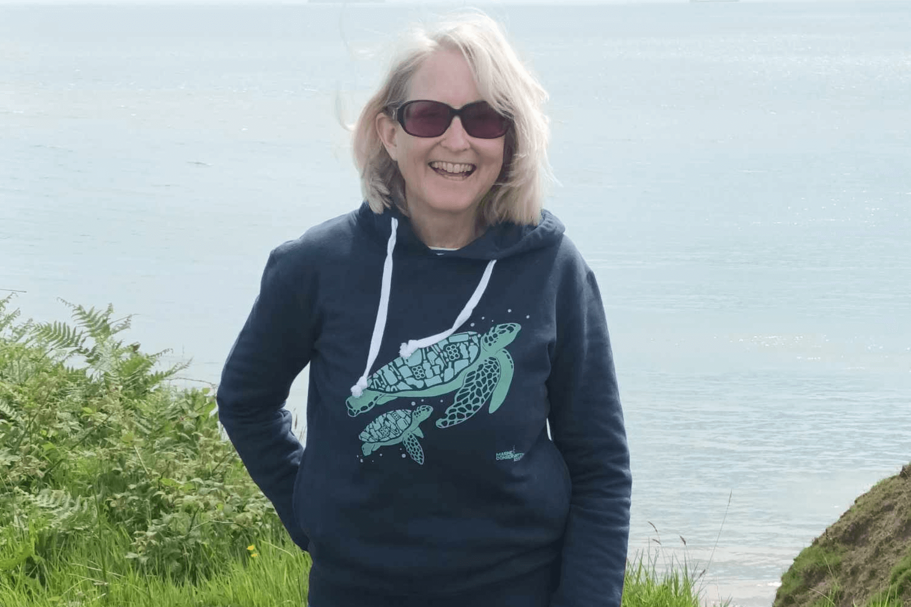 Lindsey is standing by the coast. She is wearing sunglasses and a hoodie with a turtle on it.