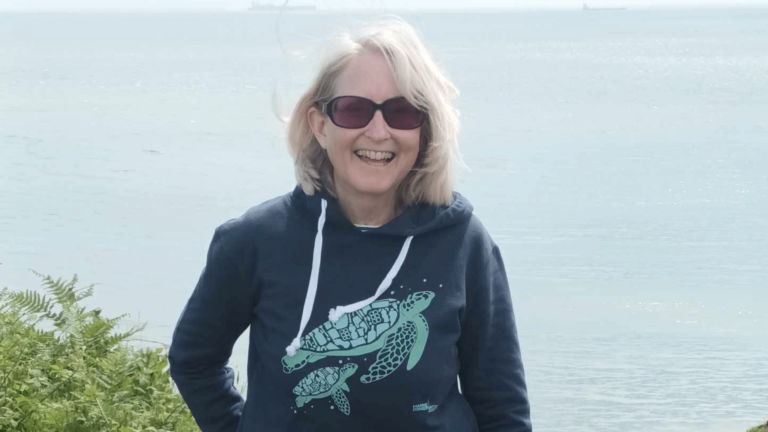 Lindsey is standing by the coast. She is wearing sunglasses and a hoodie with a turtle on it.