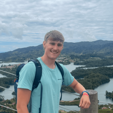 Iestyn is wearing a blue t-shirt and a rucksack. He is standing in front of hills and islands.