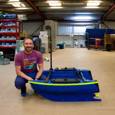 Ryan is crouching next to a Blue Boat in the Project Seagrass warehouse.