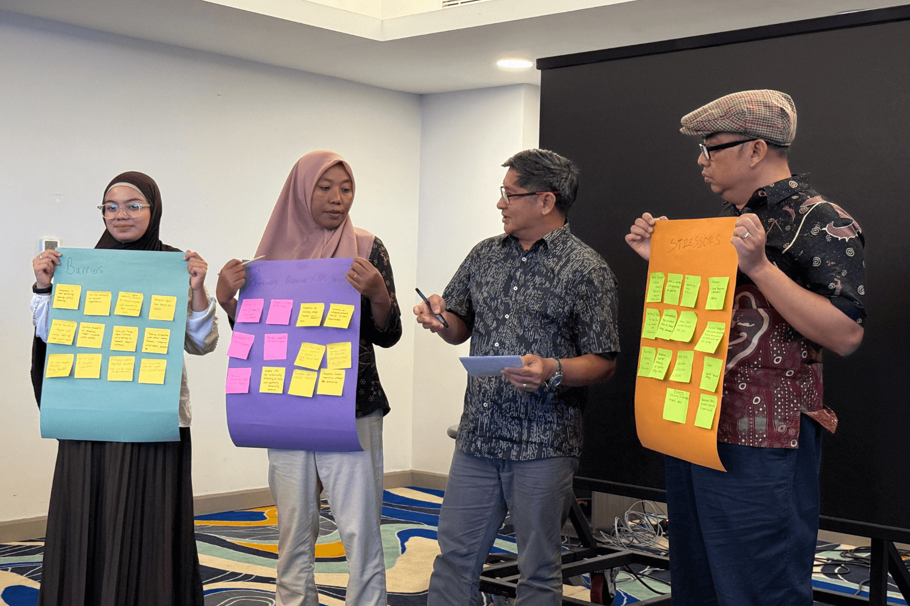 Four people are standing at the front of a room. Three members of the group are holding paper with post-it notes on as part of a workshop activity.