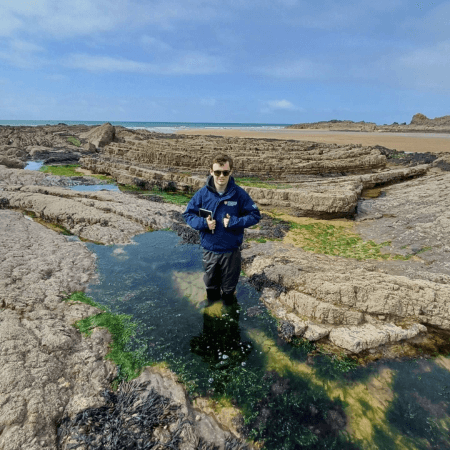William is wearing a University of Plymouth jacket and is holding a notebook. He is standing in a pool of water at the coast.