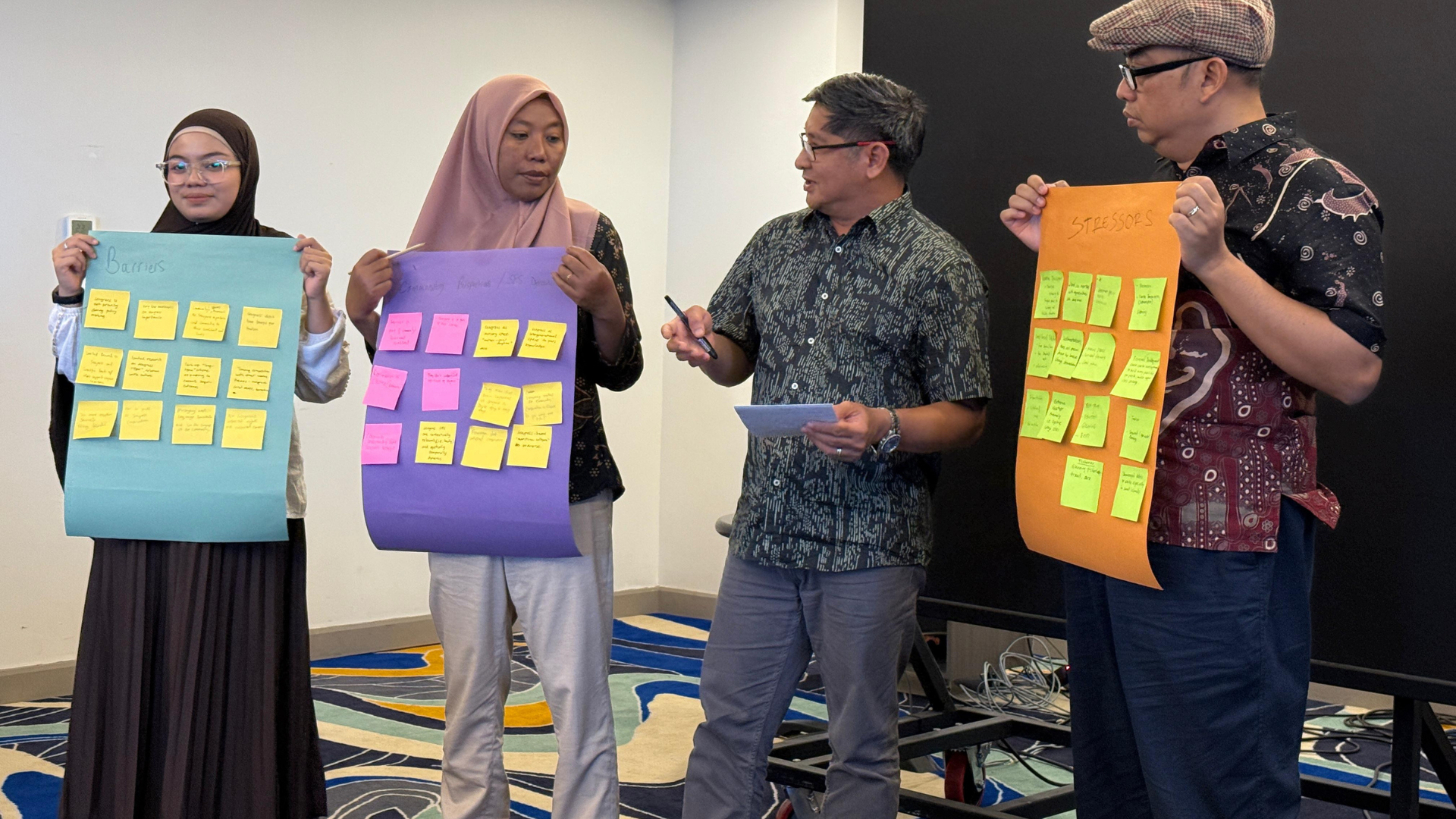 Four people are standing at the front of a room. Three members of the group are holding paper with post-it notes on as part of a workshop activity.