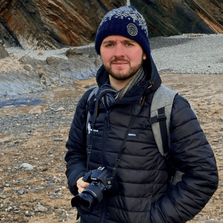 Charles is standing on a beach wearing a black Project Seagrass coat and a blue hat. He is wearing a camera around his neck.
