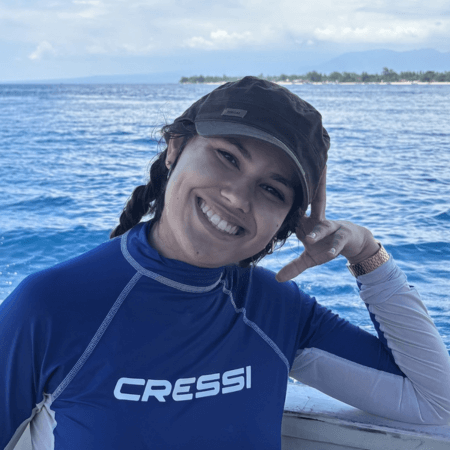 Claudia is wearing a blue and white cressi top and a brown cap. Her hair is tied back in plaits and she is leaning on one hand. The sea is in the background.