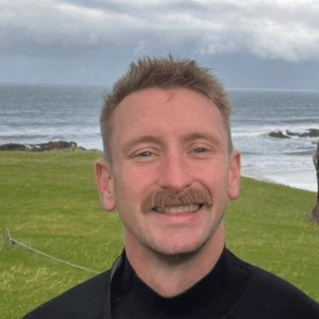 Lyle is standing at the coast with the sea behind him. He has short blonde hair and a moustache. He is wearing a wetsuit.