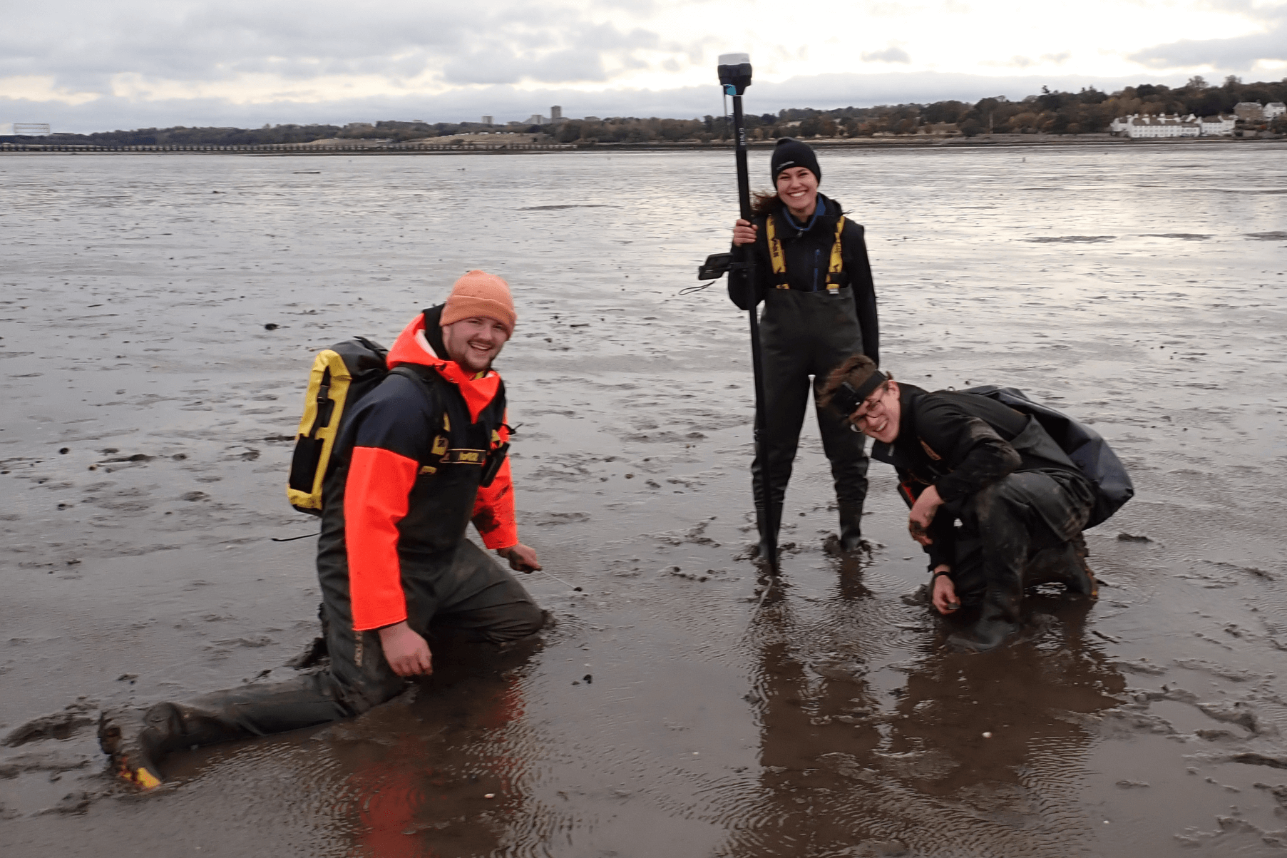 Project Seagrass interns Jasper and Ewan are out on fieldwork with fieldwork technician Claudia Foster. They are wearing waders and kneeling in the mud. Claudia is holding a DGPS to locate seagrass plots.
