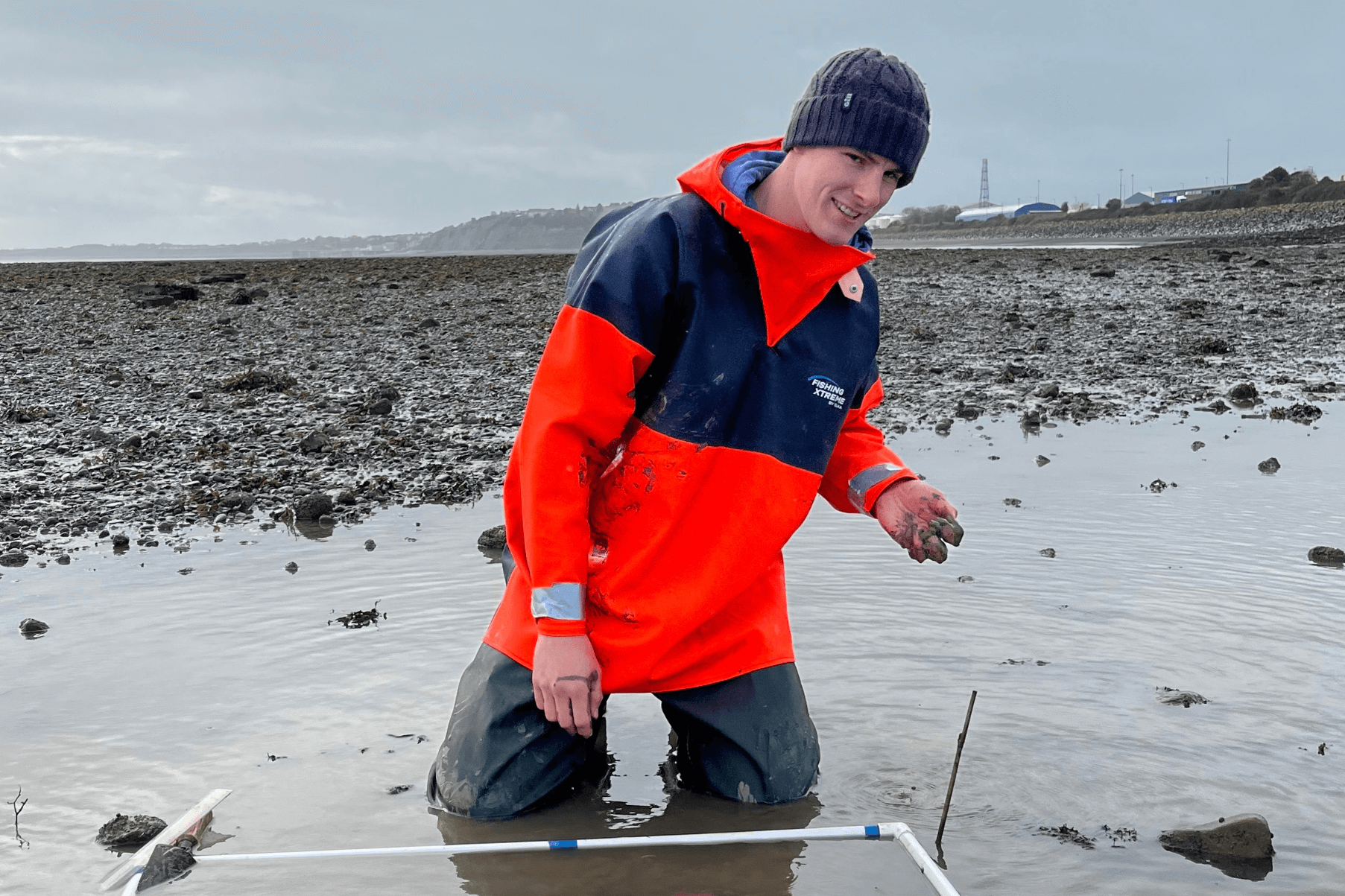 Dylan is kneeling beside a quadrat monitoring seagrass at a beach in South Wales. He is wearing a bright orange jacket and a hat,