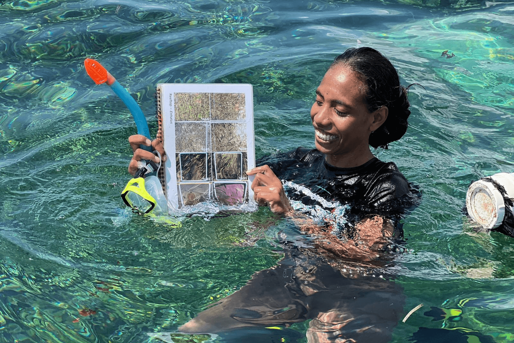 A woman is in the sea holding a seagrass monitoring training guide. She is holding a snorkel mask.