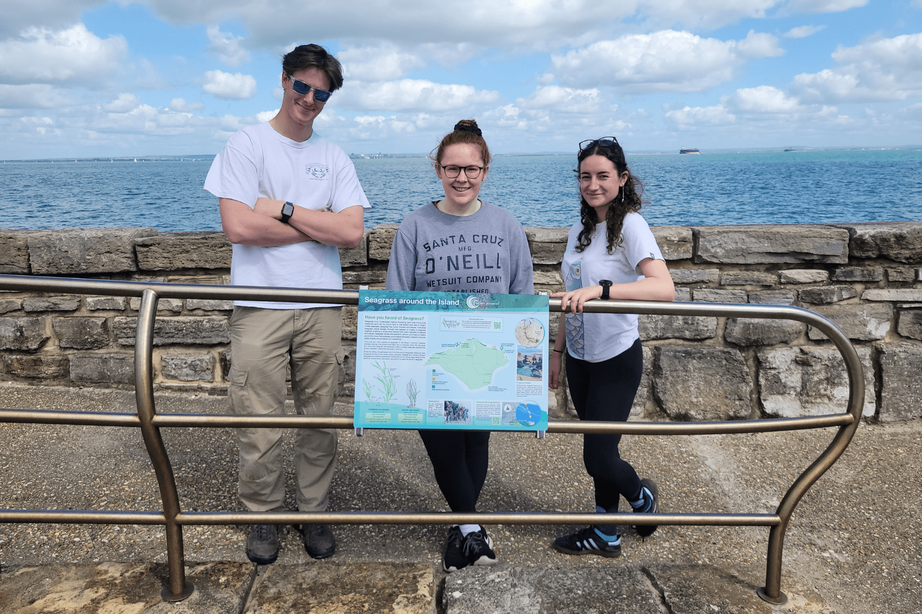 Three members of the Project Seagrass team stand behind a newly installed sign at St Helen's.