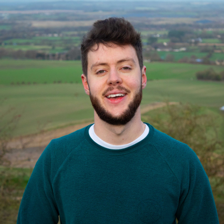 Young man with short beard and hair stands on top of a hill. Agricultural land stretches out behind him.