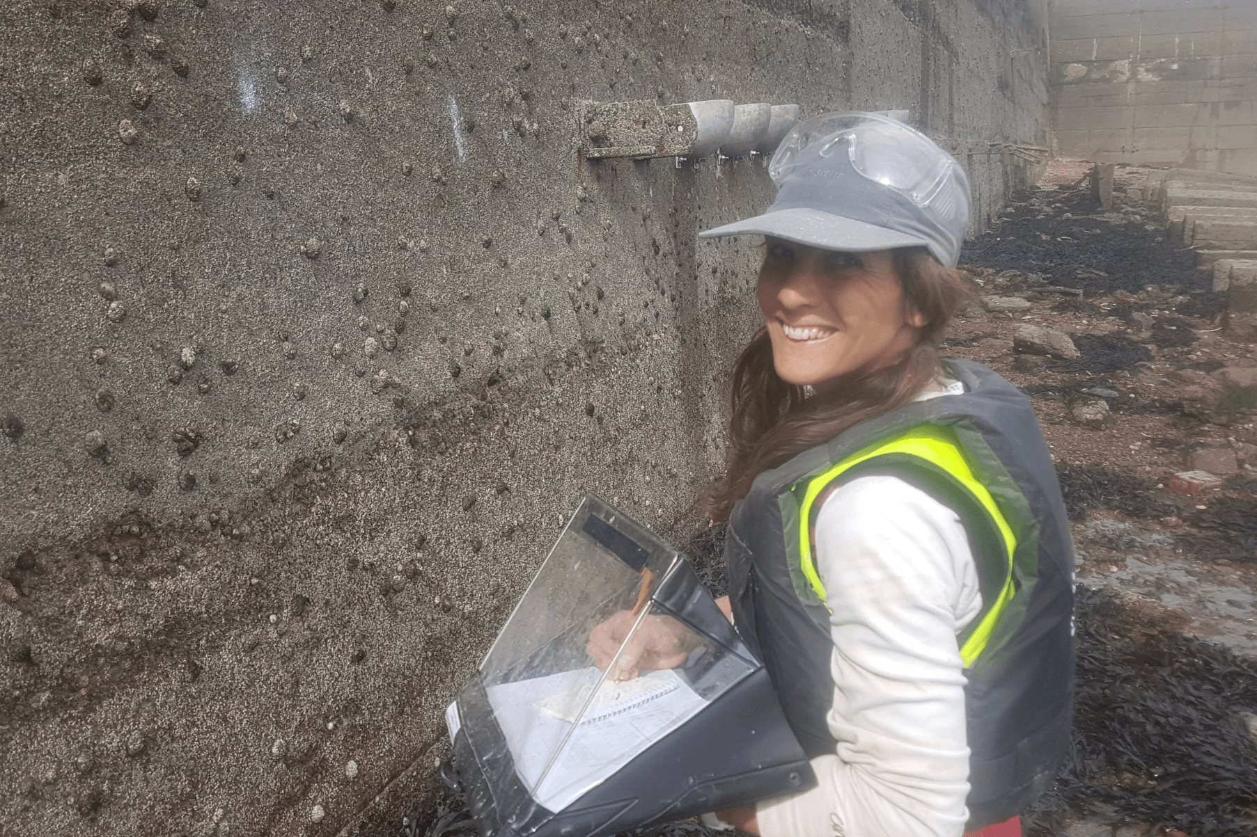Ally is crouching next to a sea wall. Shey is carrying out monitoring and making notes in a weather-writer. She is wearing a cap.
