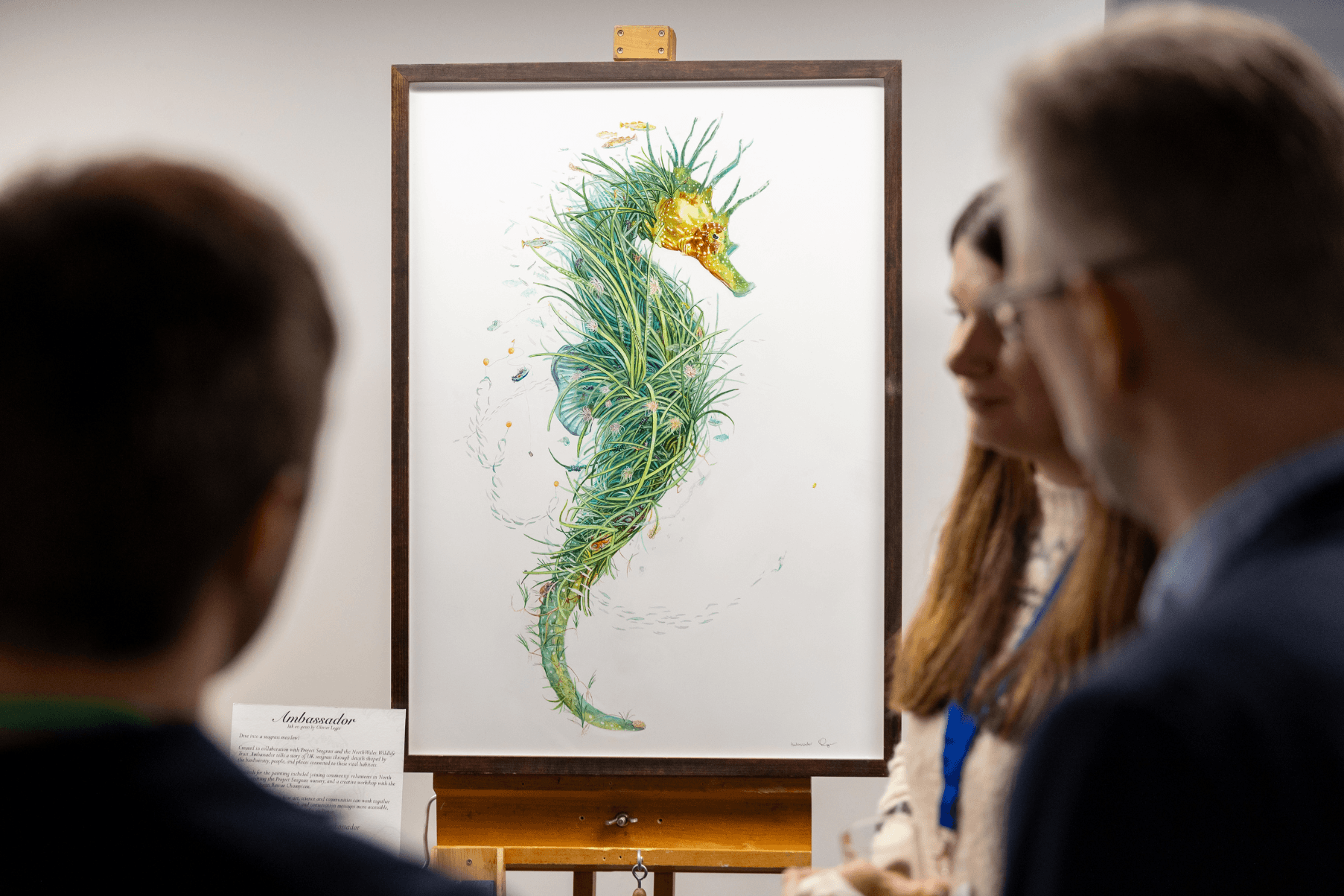 Oli's artwork is placed on a display easel at the UK Seagrass Symposium. Delegates are gathered around looking at the artwork. The artwork is of a seahorse with intricate details highlighting the importance of seagrass meadows.