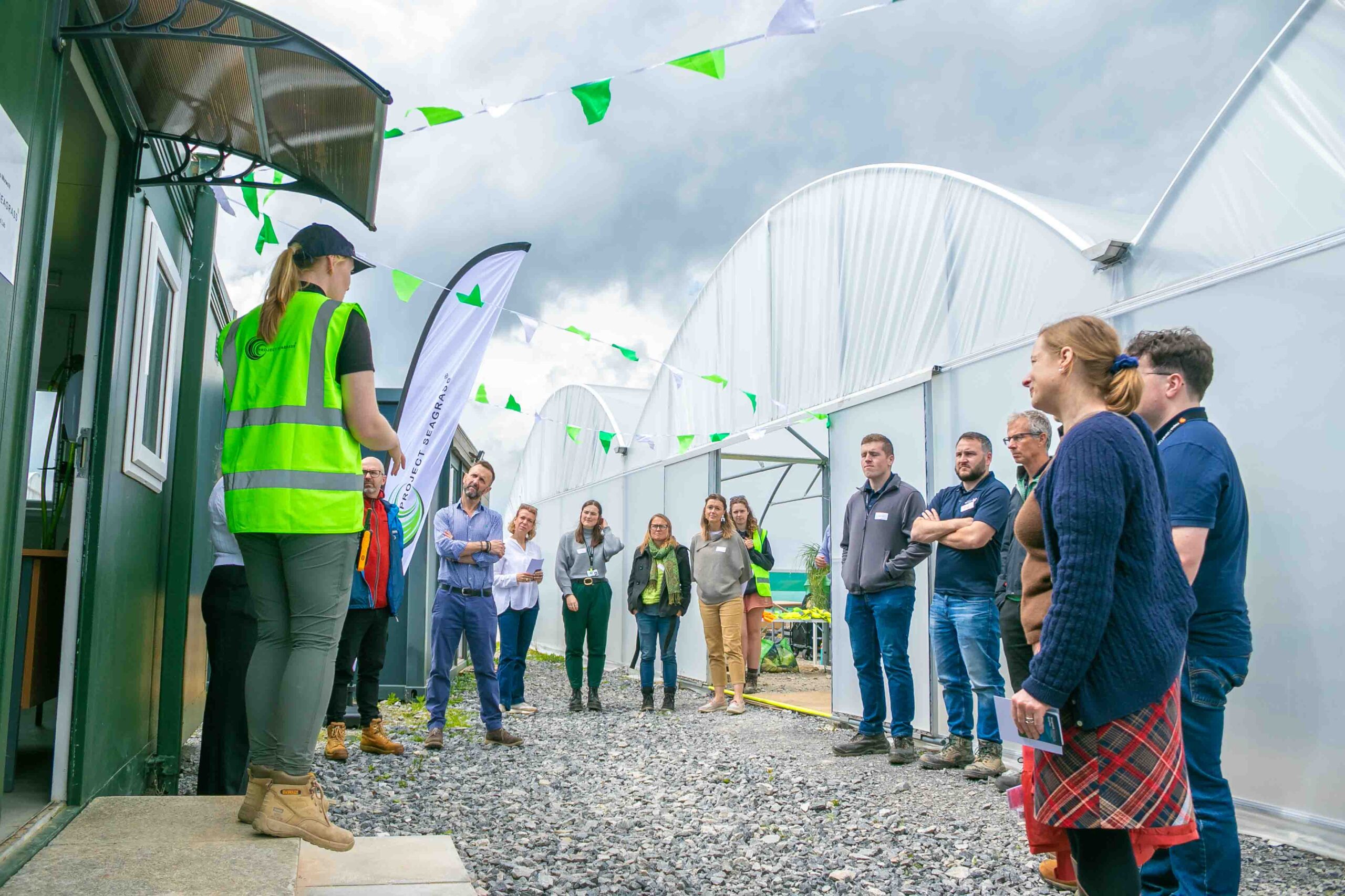 A group of people are gathered in a semi-circle around Nursery Manager Emily Yates. They are standing between a polytunnel and a portacabin which houses the seed storage facility.