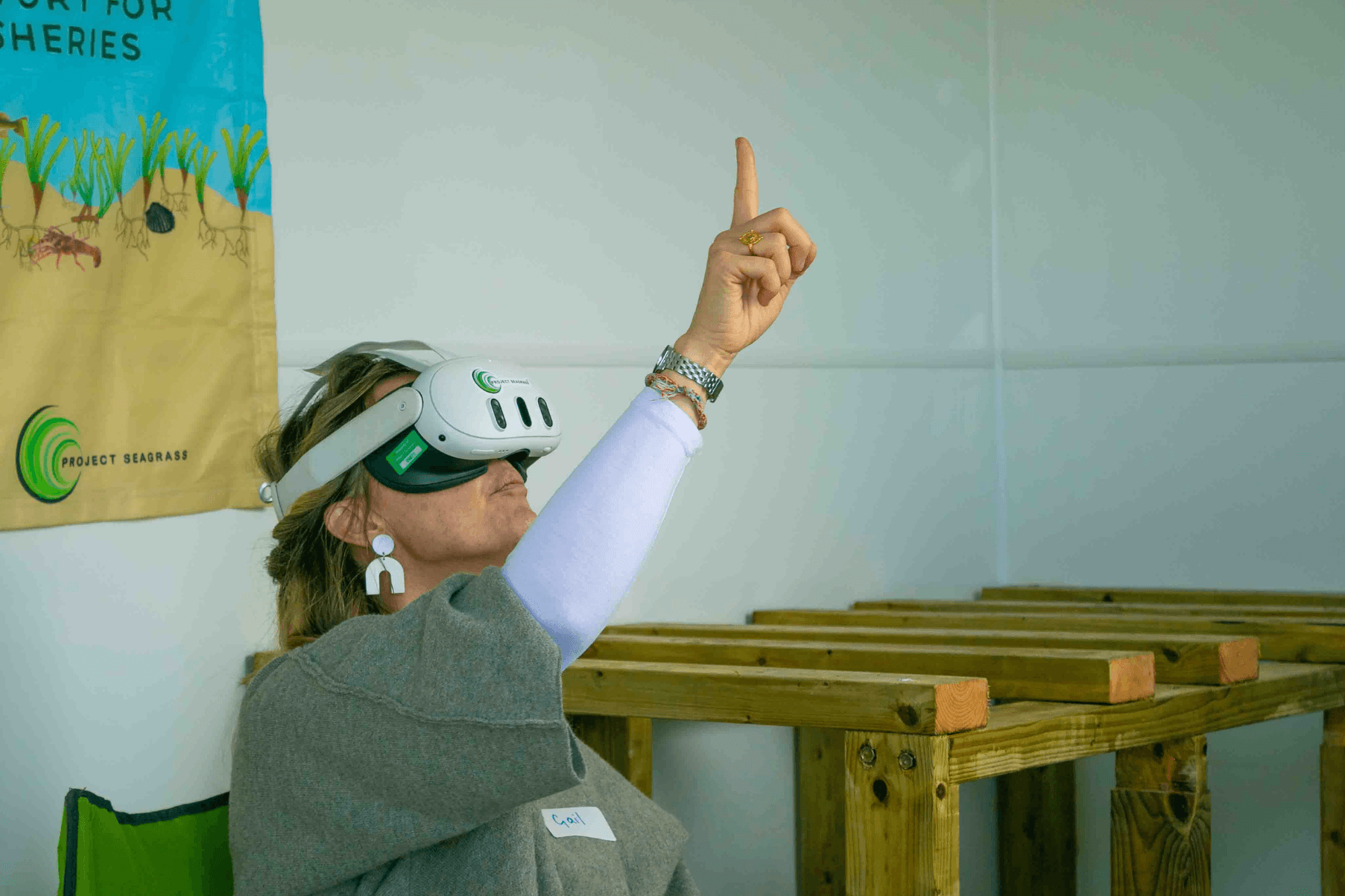 A woman is seated and wearing a VR headset. Her hand is raised as she reaches up to press a button within the experience.