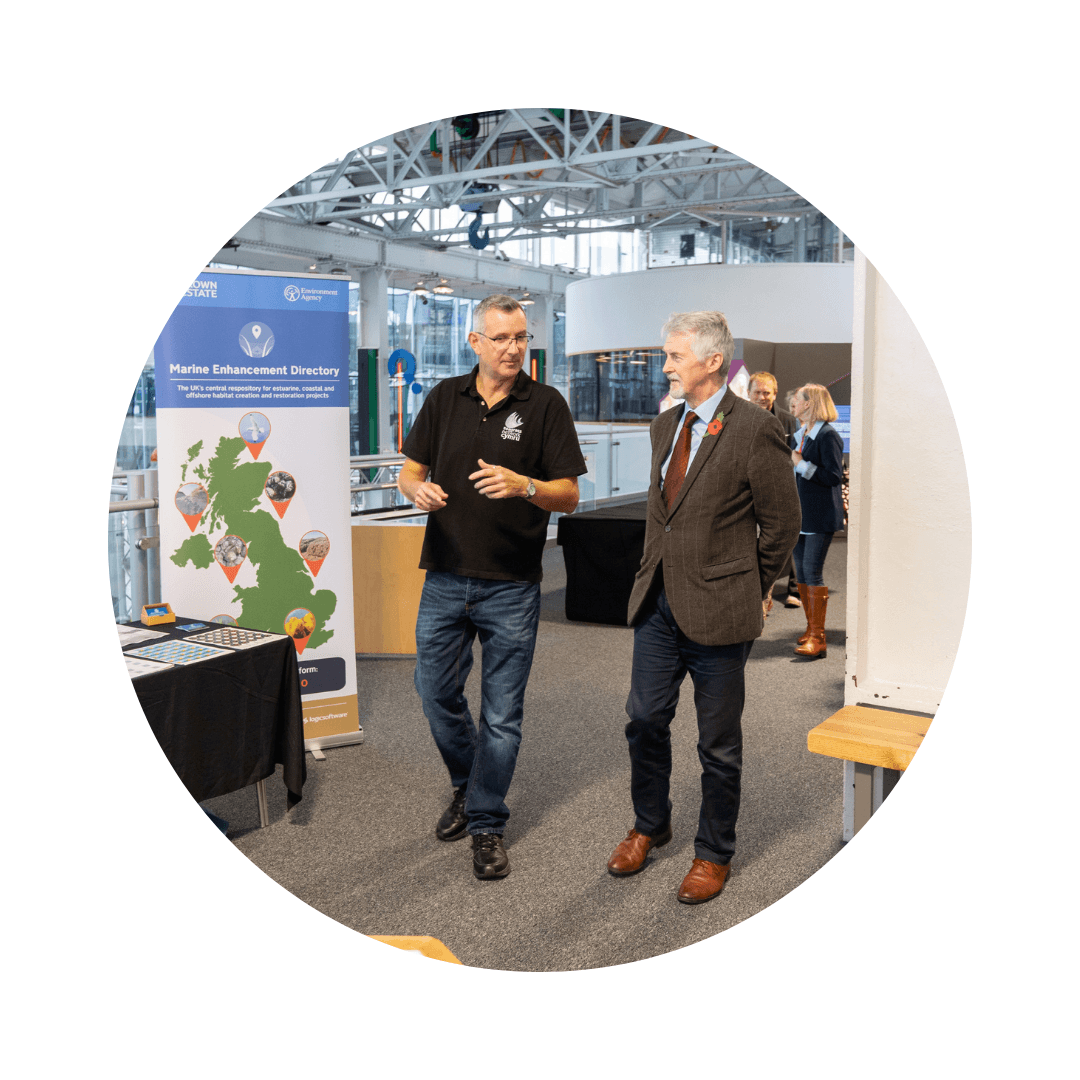 Carl is walking alongside Huw Iranca Davies. They are walking past different stands at the UK Seagrass Symposium Conference.