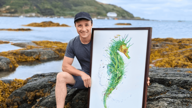 Oli is seated on a rock at the beach in Porthdinllaen, North Wales. He is holding his painting Ambassador, an artwork of a seahorse showcasing the wonders of a seagrass meadow.