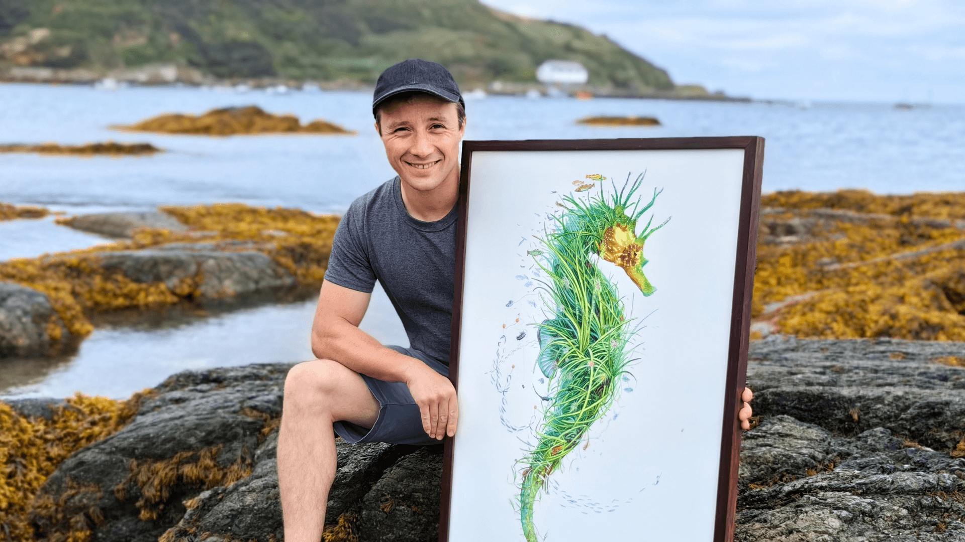 Oli is seated on a rock at the beach in Porthdinllaen, North Wales. He is holding his painting Ambassador, an artwork of a seahorse showcasing the wonders of a seagrass meadow.