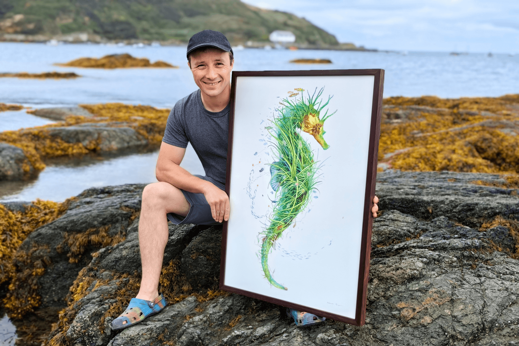 Oli is seated on a rock at the beach in Porthdinllaen, North Wales. He is holding his painting Ambassador, an artwork of a seahorse showcasing the wonders of a seagrass meadow.