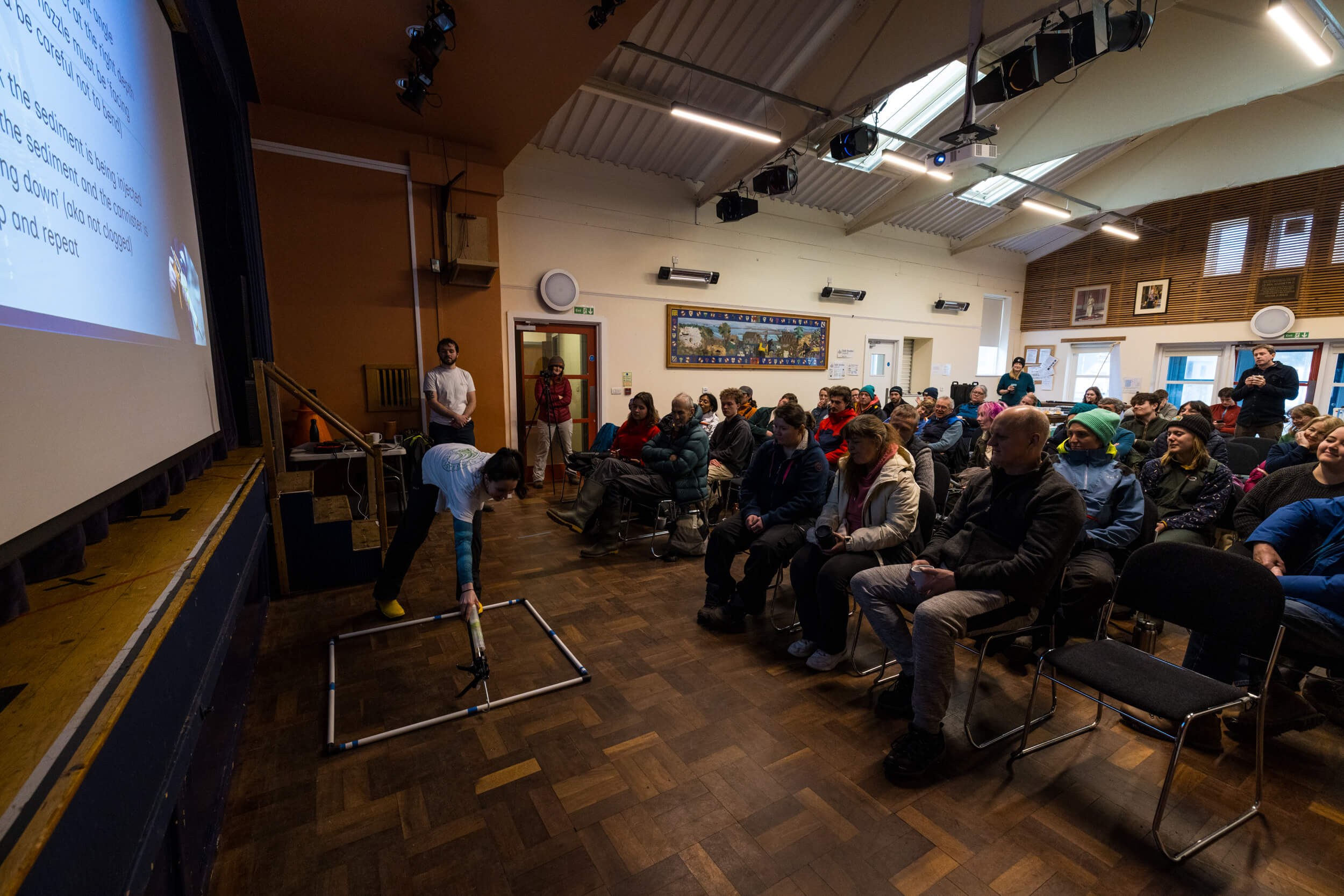 A hall full of people are seated watching a demonstration of how to plant seagrass seeds.