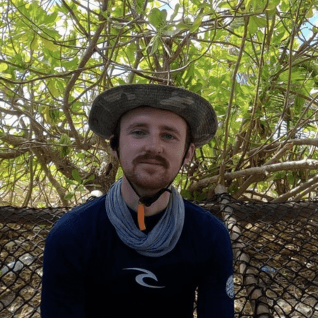 Matthew is sitting in front of a mangrove. He is weating a hat.