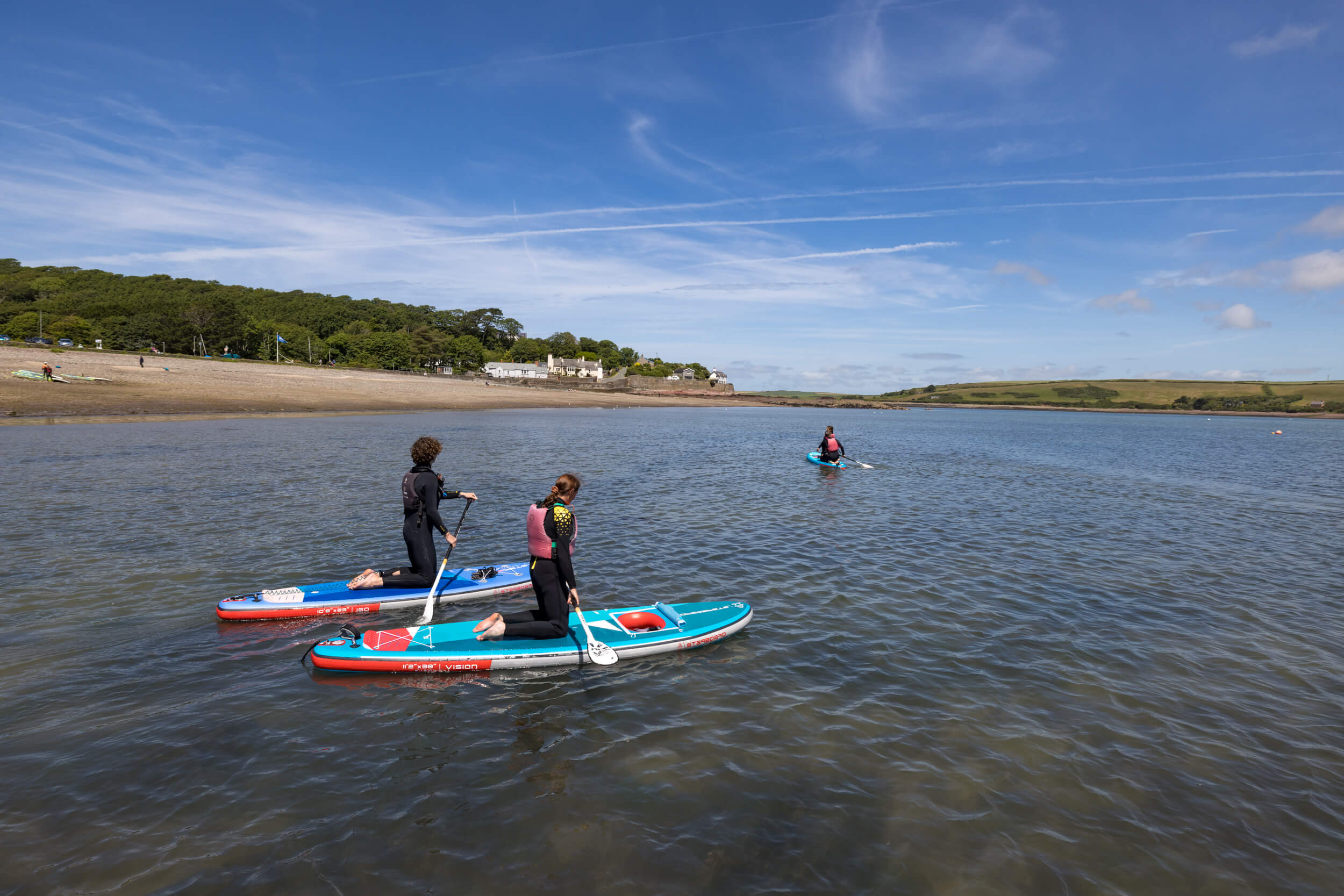 Three paddleboarders explore the seagrass in Dale Bay, Pembrokeshire. They are wearing wetsuits and lifejackets. The sky is blue.