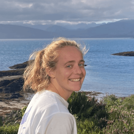 Issy is standing by the sea. She is wearing a white t-shirt and looking over her shoulder towards the camera.