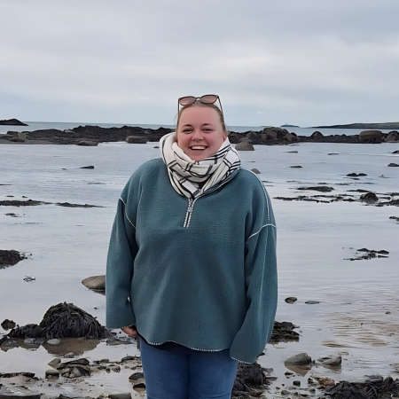 A woman facing the camera with blonde hair stands at medium distance on a sandy beach with rocks and bladderwrack at low tide.