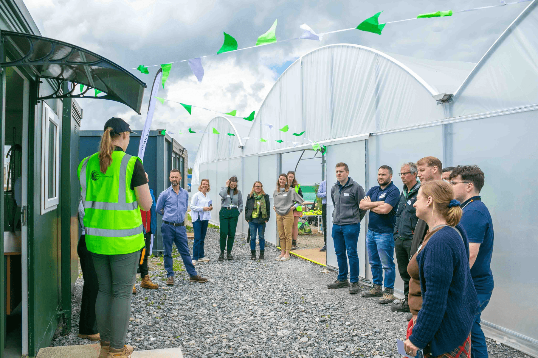 Nursery Manager Emily is wearing a high vis jacket and standing in front of a semi-circle of people visiting the Seagrass Nursery. The large triple span polytunnel is adorned with bunting.