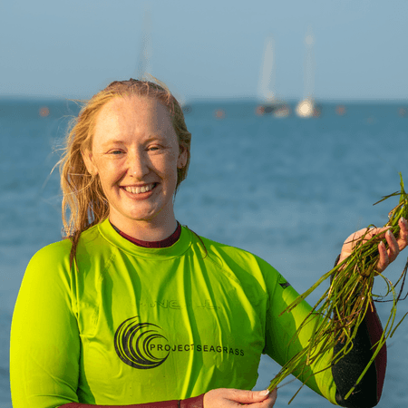 Emily is wearing a bright yellow rash vest with the Project Seagrass logo on in and is holding seagrass seeds collected in the seagrass meadow at Yarmouth