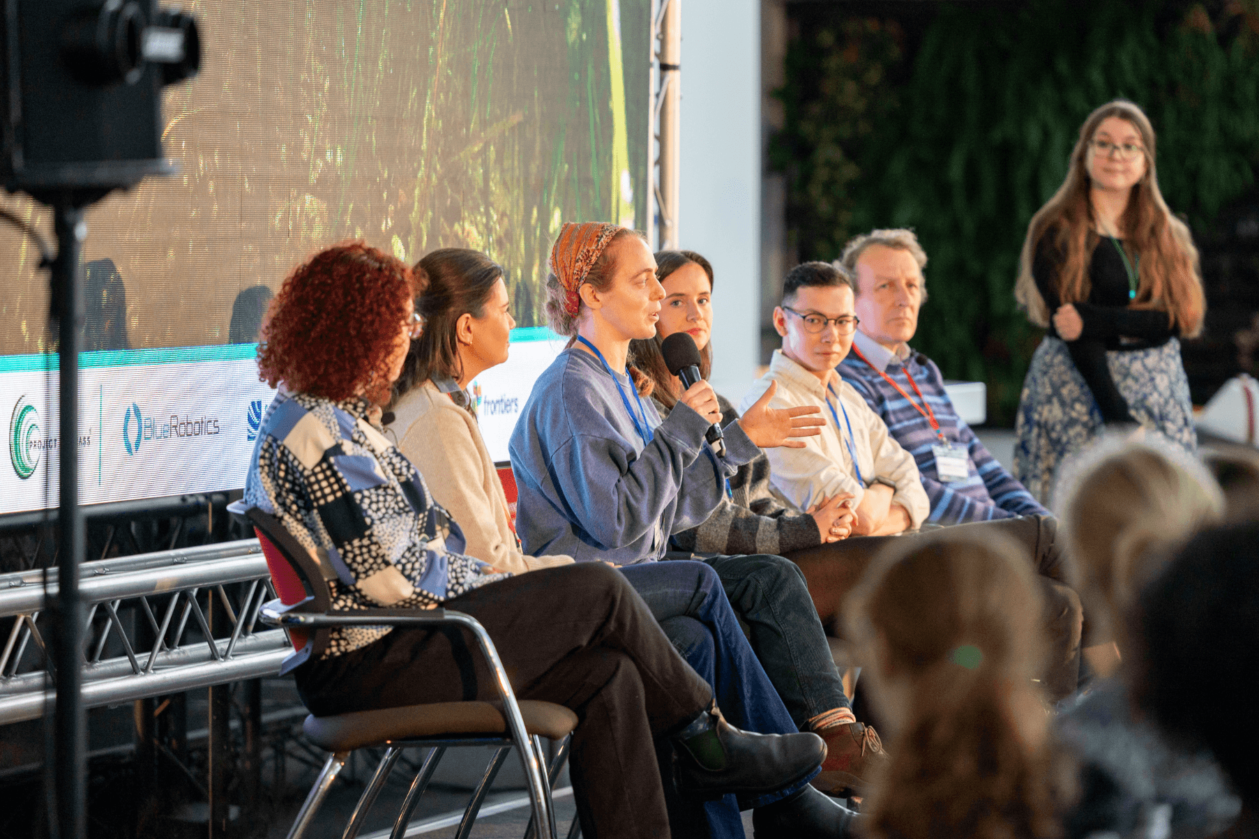 5 scientists are seated on a stage in front of a screen as part of a panel discussion at the UK Seagrass Symposium. One of the speakers holds a mic and responds to a question from the audience. To the side, the session host stands by a podium.