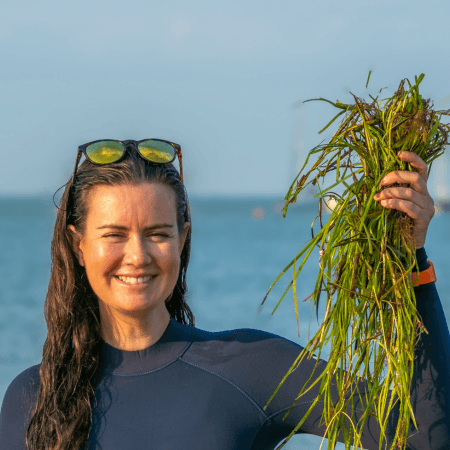 Anouska is standing at the edge of the water wearing a wetsuit. She is holding a large handful of seagrass, collected as part a seagrass seed collection. She has sunglasses on her head.