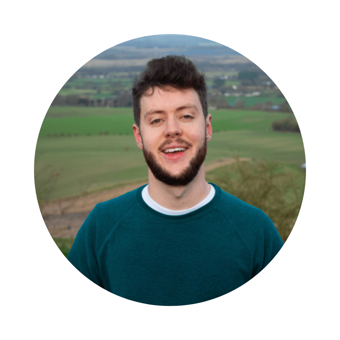 Young man with short beard and hair stands on top of a hill. Agricultural land stretches out behind him.