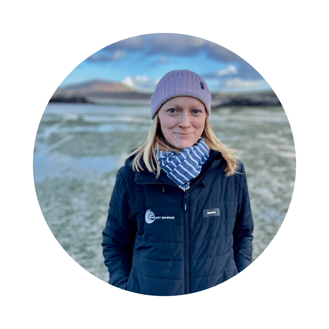 Leanne is wearing a black Project Seagrass jacket, a purple hat and a blue and white striped scarf. She is standing with her hands in her pockets by a seagrass meadow.