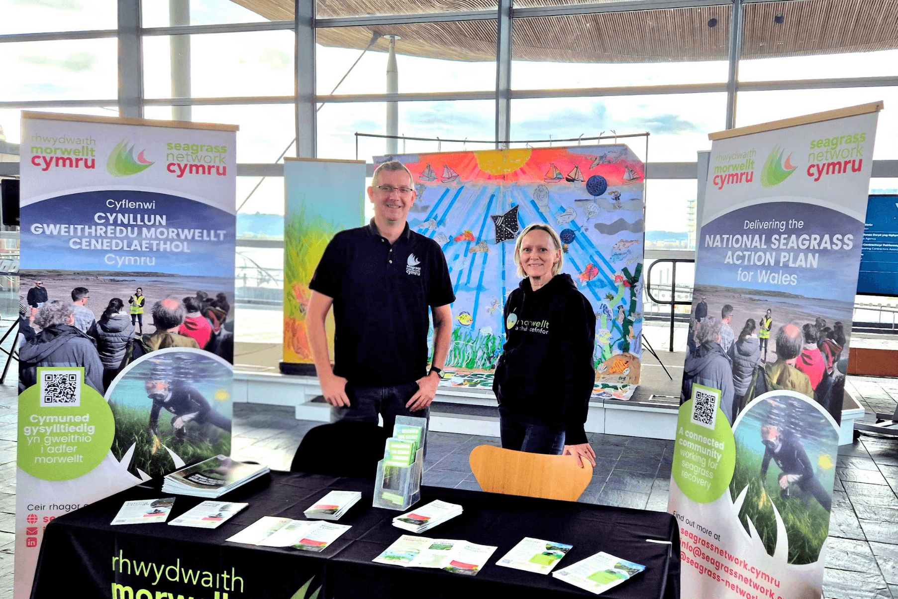 Leanne and Carl stand behind a table with information about Seagrass Network Cymru on it. Two banners are either side of the table. In the background a large artwork called the Meadow of Hope displays artwork and messages for the ocean written by schoolchildren.