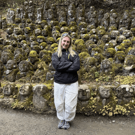 Laura is standing in front of a stone walls with faces carved into it. She is wearing a black jacket and white trousers and has her arms folded. She has blonde hair and a pair of sunglasses on her forehead.