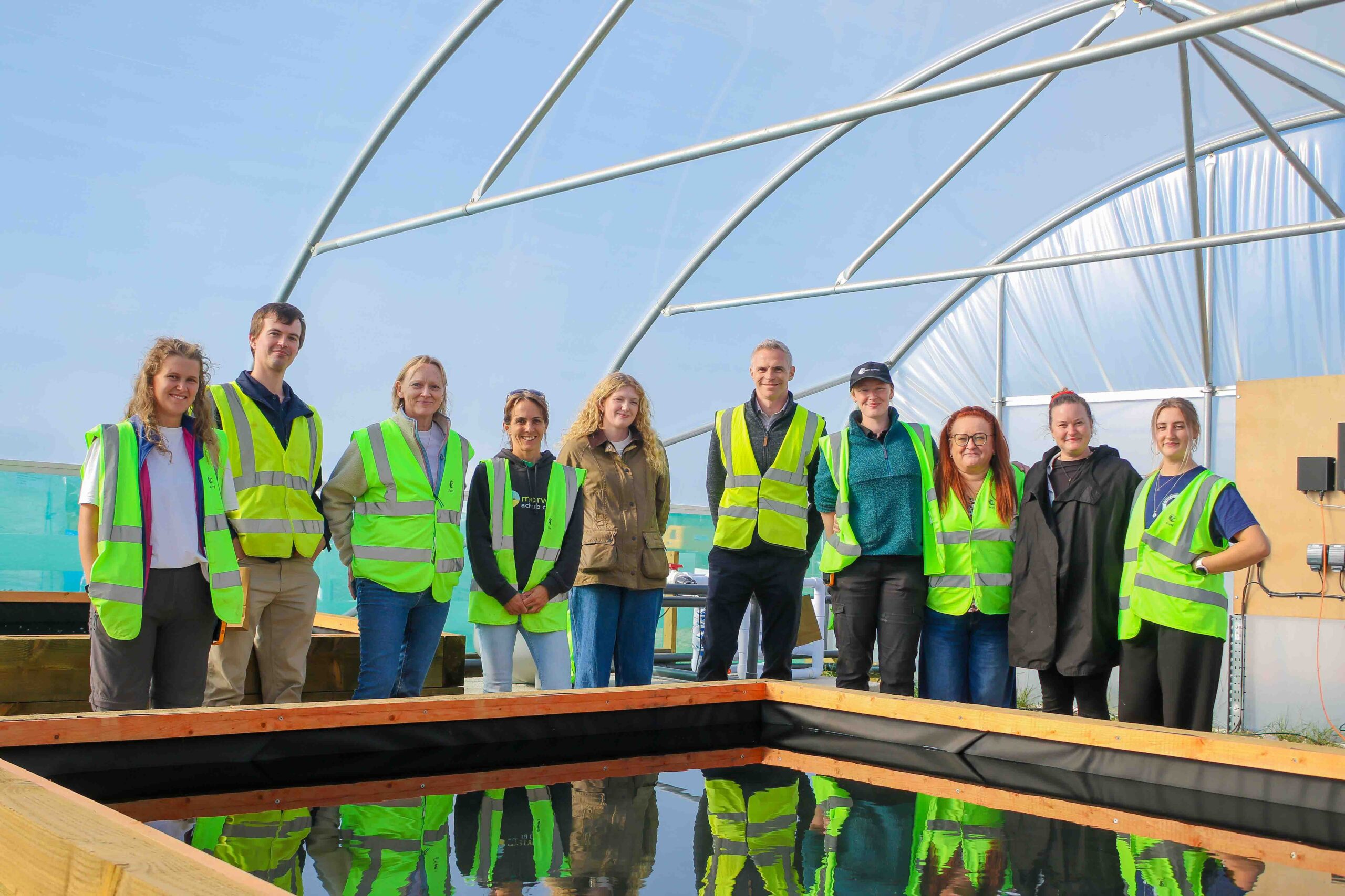 Project Seagrass team members and funders and partners are gathered around one of the ponds in the new polytunnel. They are wearing high vis vest following a tour of the site.