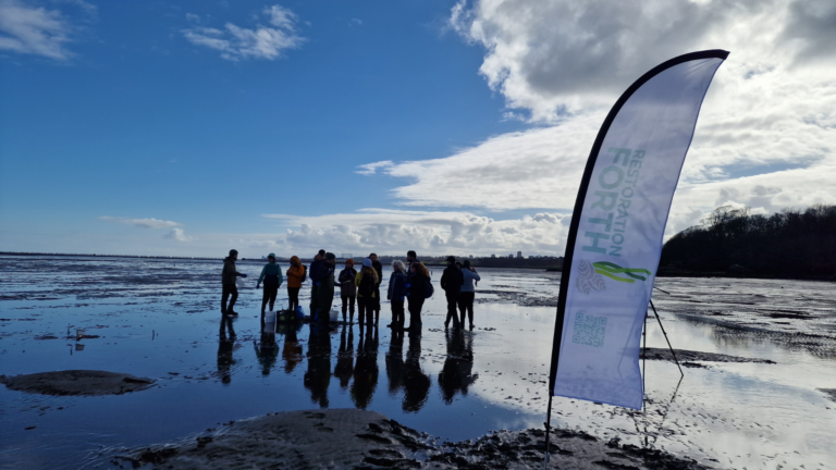 A banner flag with the Restoration Forth logo is set up on the beach. A group of staff and volunteers are gathered next to it. The sky is blue with some clouds.